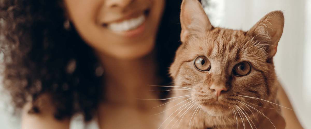 A smiling woman with curly hair holds an orange tabby cat close to her face. The cat, sharp in focus, looks directly at the camera while the woman smiles warmly in the background.