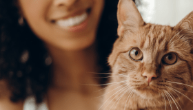 A smiling woman with curly hair holds an orange tabby cat close to her face. The cat, sharp in focus, looks directly at the camera while the woman smiles warmly in the background.